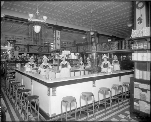 Interior view of Dutch Mill Restaurant, 1541 Champa Street, Denver, Colorado; shows a man, a woman, and waitresses, behind a marble counter and metal barstools, in Dutch costume; they wear hats, dresses, collars, neck bows and aprons. A coffee machine, glassware, and menus are by containers of straws. Merchandise includes dishcloths, "Calarai Figs," cans and jars. Christmas garland and poinsettias decorate all including second floor balustrades.