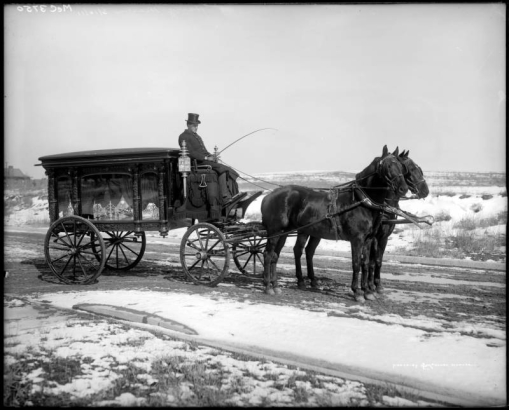 View of a horse-drawn hearse on a muddy road, probably in Denver, Colorado; the driver wears a top hat and coat. Snow covers fields.