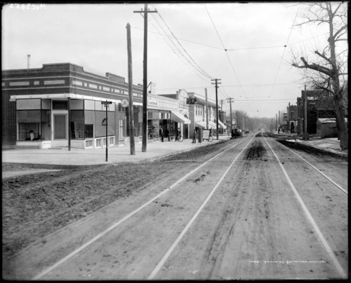 View of 32nd (thirty-second) Avenue in Denver, Colorado; shows brick commercial buildings, streetcar railway tracks, intersection of Mead Street, horse-drawn wagon, an automobile, and signs: "Long Distance Telephone," "Millinery," "Denver Bible Institute," "Tin Shop and Hardware,"  "Grocery," "Highland Theater," "5-cent," "Bakery," and sandwich board:  "Spuds and Homemade Cakes."