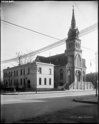 View of Sacred Heart Catholic Church at 28th (Twenty-eighth) and Larimer Streets, in Denver, Colorado, with corbeled brick, intersecting gables, central stepped steeple topped with a cross, stairs, and gothic stained glass windows. An apartment building, iron fence, and mailbox are on the corner.