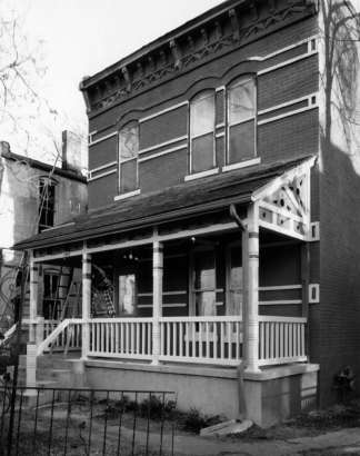 View of 2754 Stout Street in the Curtis Park neighborhood (Five Points) in Denver, Colorado; shows a brick house with corbeling and brackets; a man works on the covered porch.