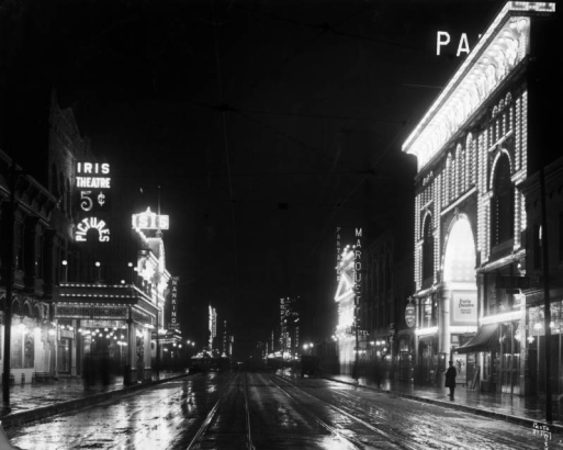 View of Curtis Street, in Denver, Colorado; shows wet pavement, and electric signs: "Iris Theatre 5 cent Pictures," "Isis," "The Nanking Chop Suey," "Princess," "St. James," "Tabor Grand," "Marquette Hotel," "Pantages," and "Paris Theater."