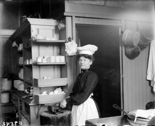 Interior view of a Daniels and Fisher store kitchen, in Denver, Colorado; shows a woman in a chef's hat with food. Shelves hold dinnerware and crockery.