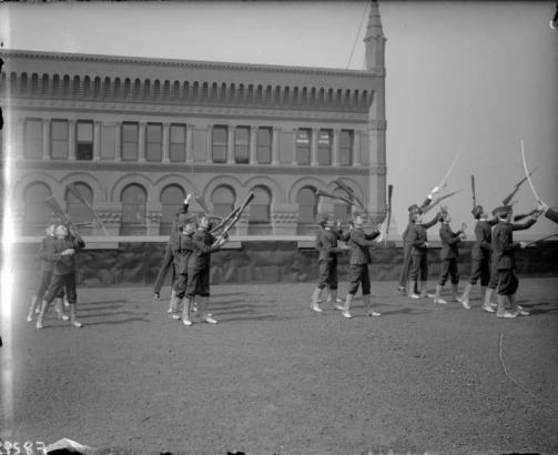 View of Daniels and Fisher staff, in Denver, Colorado; shows men and boys in uniform, in formation, holding rifles and swords. The Continental Building is in the background.