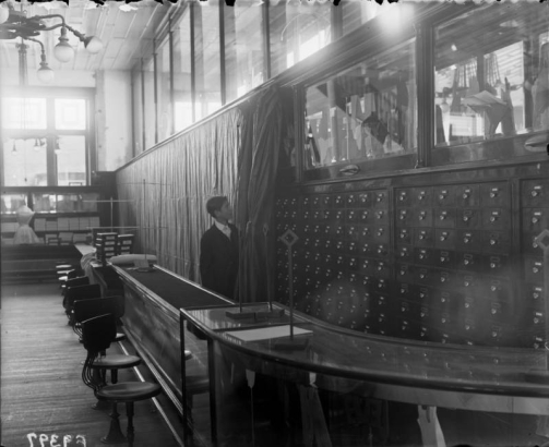 Interior view of the Daniels and Fisher store, in Denver, Colorado; shows glass counters and display cases. A boy draws curtains by shelves of drawers.