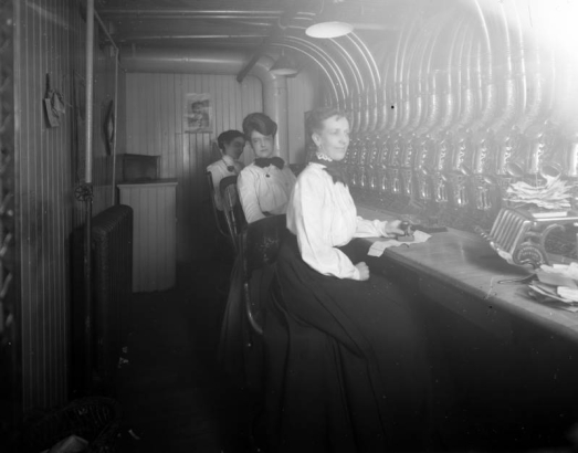 Interior view of a Daniels and Fisher store in Denver, Colorado; shows women seated at a pneumatic messaging device; one holds a rubber stamp by a "Staats Money Changer" machine.