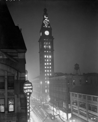 Night view of the Daniels and Fisher Stores tower, in Denver, Colorado; shows 16th (Sixteenth) Street with Christmas decorations, and neon signs: "Harry Huffman's Tabor Musical," and "D & F (Santa Claus)."