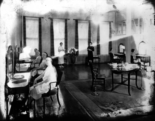Interior view of the Daniels and Fisher Stores Company, in Denver, Colorado; shows women at tables with mirrors, cosmetics, flowers, and an ash tray.