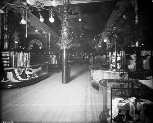 Interior view of the Daniels and Fisher store in Denver, Colorado; shows glass display cases of lace, and arrangements of pine boughs.
