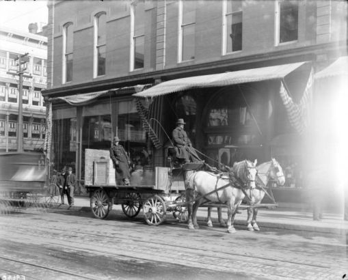 View of a Daniels and Fisher store delivery wagon and horses in Denver, Colorado; shows men and a teenaged boy in bowler hats. Storefronts are in the background.