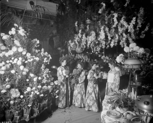 Interior view of the Daniels and Fisher store in Denver, Colorado; shows women in brocade kimonos holding chrysanthemums and pouring tea. Lamps, a plant, and dishes are on a table.