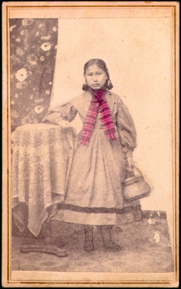 Studio portrait (standing) of a Native American (Arapaho) girl. She holds a basket and rests her arm on a table. She wears a dress and has a scarf around her neck.