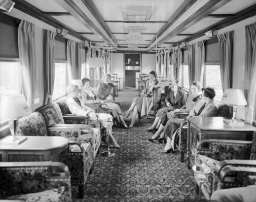 Women in high heels, dresses, skirts and hats pose on furniture inside the Denver and Rio Grande Western Railroad dining lounge car refurbished from an ex-Western Pacific dining car, possibly parked in Denver railroad yard. Interior decorated with Art Deco arm chairs, sofas, tables, lamps, smoking stands and ornate metal wall partitions. Upholstery features floral and geometric motifs. Possibly taken during the Summer of 1939.
