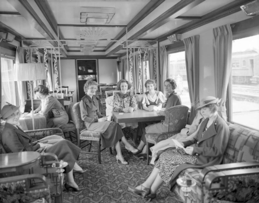 Women in high heels, dresses, skirts and hats pose on furniture inside a Denver and Rio Grande Western Railroad dining lounge car, refurbished from a Western Pacific dining car, parked possibly in the Denver, Colorado railroad yards. Some of the women play cards. Interior design is Art Deco with arm chairs, sofas, tables, lamps, smoking stands and ornate metal wall partitions. Upholstery features floral and geometric motifs. Possibly taken during the Summer of 1939.