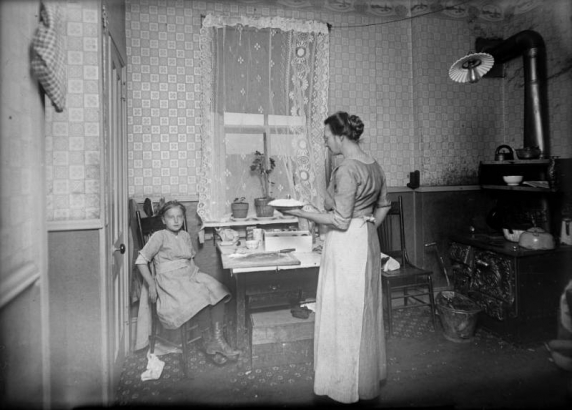 Interior view of a kitchen in Denver, Colorado; shows a girl, a stove, a cutting board, and a woman holding a pie.