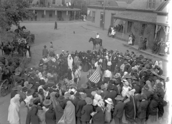 Taos Pueblo Indians and tourists form a circle around dancers with a United States flag in the Taos Plaza, Taos, New Mexico. Native Americans (Taos Pueblo) are wrapped in blankets, the sightseers wear hats. Onlookers stand on porches or portals of enclosing buildings. A sign reads: "Post Card Views, Indian Curios, Kodak Finishing."