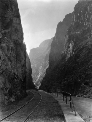 View along the Arkansas River and the Denver and Rio Grande Western railroad tracks, with the Hanging Bridge in distance, Royal Gorge, Fremont County, Colorado. Wooden Canon City water supply pipeline shows tunneling in and along the steep granite wall along river.