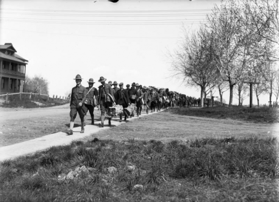 A long line of troops, some in uniform, others in civilian clothes, march along a walkway next to the barracks, Fort Logan, Colorado, a World War I assembly point for young soldiers. Some carry Army musette bags.