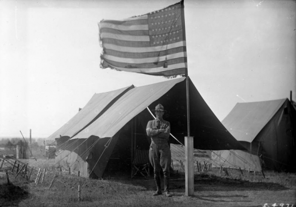 A soldier, in uniform, poses outside a staked walled tent beside a flag pole with a fringed American flag at Fort Logan, Colorado.