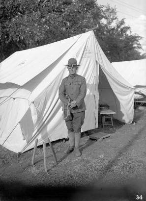 The bugler poses outside a Schaeffer tent in a uniform consisting of a jacket with two breast pockets with one bar above the right one, jodhpurs, boots, canvas gaiters and a wide brim hat with Montana creases, Loretto Heights service camp, Denver, Colorado.