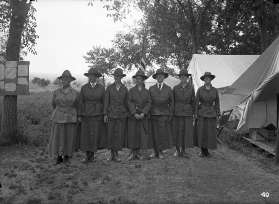 Female instructors pose in uniform beside tents and a United States flag, Loretto Heights service camp, a WWI training center for women, Denver, Colorado. One woman holds a baton.