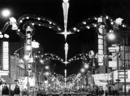 Nighttime view of 16th (Sixteenth) Street in downtown Denver, Colorado. Automobiles drive under Christmas lights and decorations that span the street. Signs read: "Denver," "Baurs," "Douglas Optical," and "Paramount."