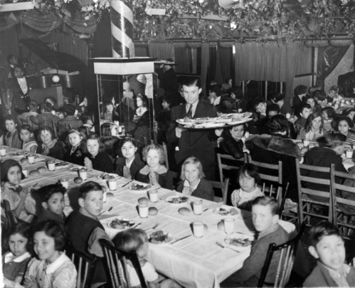 Boys and girls (white, Hispanic, African American, and Italian American) prepare to eat a Thanksgiving dinner in the Tivoli Terrace nightclub at 1900 West 32nd (Thirty-second) Avenue in the Highland neighborhood of Denver, Colorado. A waiter carries a large tray of dinner plates between tables.  A Black woman singer and pianist prepare to entertain.