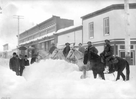 View of a heavy snowfall on Sixth (6th, Alpine) Street in Georgetown, Colorado. The Spruance Building and Kneisel-Curtis-Seifeld building are on the south side of the street. Five men ride large horses; five men stand (wearing heavy coats and hats) surrounded by piles of snow.