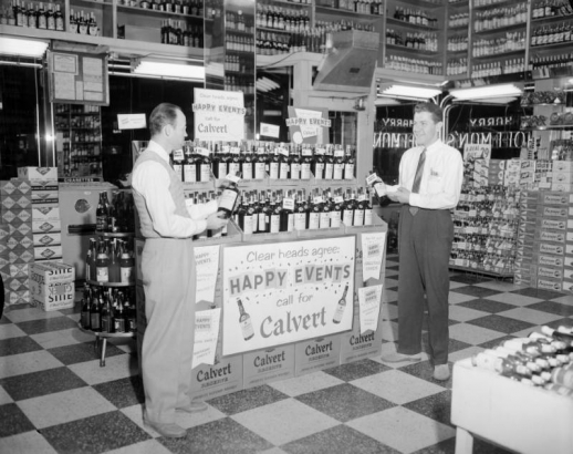 Interior of Harry Hoffman's Cut Price Liquor store at night in Denver, Colorado. Two men (George Papadias on the right) hold bottles of whisky and stand by advertising.