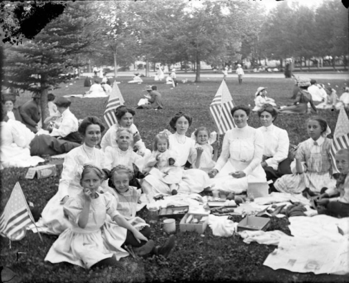 A group portrait of women and girls as they picnic outdoors possibly in City Park, Denver, Colorado. A few girls hold U. S. flags. Other groups of people have picnics nearby.