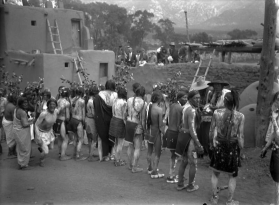 View of Native American (Taos Pueblo) men at Taos Pueblo, New Mexico, wearing moccasins, breechcloths, and body paint, during a festival. They carry branches; adobe pueblo dwellings and white spectators are in the background.