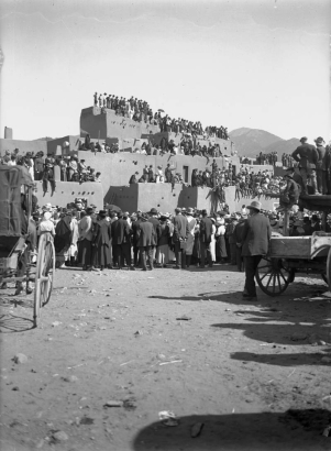 View of Taos Pueblo, New Mexico, during the San Geronimo Day festival; shows a crowd of spectators including Native Americans (Taos Pueblo) and whites. Many sit on adobe pueblo dwellings.