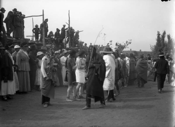 View of the San Geronimo festival at Taos Pueblo, New Mexico; shows Native American (Taos Pueblo) men wearing body paint and holding branches. White men and women watch from the side.