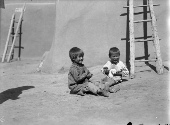 Portrait of Native American (Taos Pueblo) boys by a ladder and adobe walls at Taos Pueblo, New Mexico; they play with a puppy.