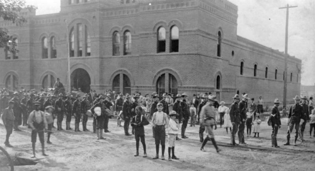 Colorado National Guard, mustered to combat the labor strike of the Western Federation of Miners, march on Curtis Street in front of the brick Armory building at 2565 Curtis, Denver, Colorado. A crowd, including two Black children, stand and watch.