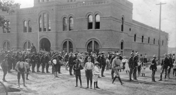 Colorado National Guard, mustered to combat the labor strike of the Western Federation of Miners, march on Curtis Street in front of the brick Armory building at 2565 Curtis, Denver, Colorado. A crowd, including two Black children, stand and watch.