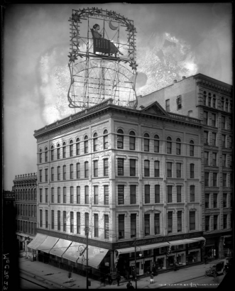 View of the Davis & Shaw Furniture Co. in the Pioneer Building at the corner of 15th (Fifteenth) and Larimer Streets, Denver, Colorado; shows ornate, electric-light advertising sign on roof, James Petersen Shoe Shop, and Ibbs Barber Shop at right; pedestrian traffic, parked cars.
