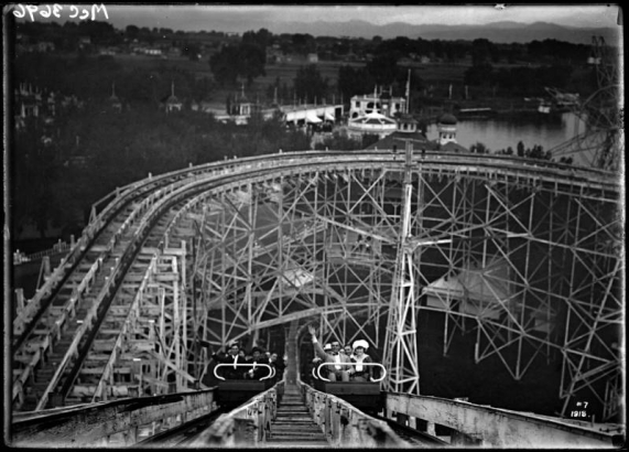 View of riders on a roller-coaster at White City amusement park (later called Lakeside) west of Denver, Colorado; view taken from top of incline looking down at two car-loads of waving riders.