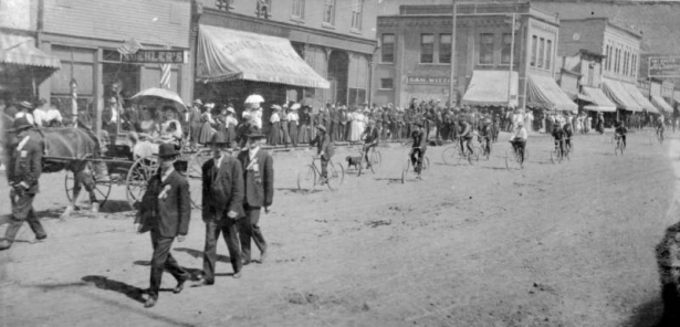 Men march and ride bicycles in a parade in downtown Denver, Colorado. Women ride in a horse-drawn wagon as part of the procession. Signs on buildings read: "Koehler's," "Crooke & Fuller Mine & Mill Supplies," and "Sam Wittow."