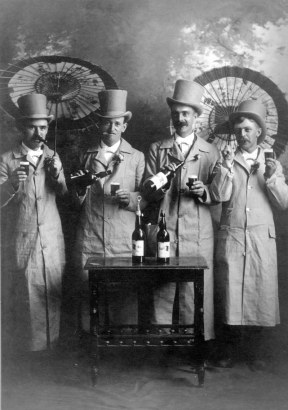 Studio portrait of Charlie Nelson, Thomas C. Browne, Douglas Patilla, and Charles Smith in Denver, Colorado. The men hold parasols and pour beer from bottles. They wear top hats and overcoats.
