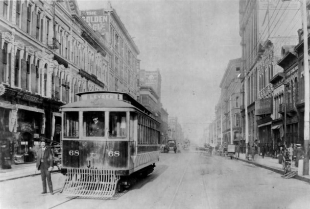 A streetcar conductor and a streetcar operator pose near and in Denver City Tramway Company car number 68 on Lawrence Street between 16th (Sixteenth) and 17th (Seventeenth) Streets in downtown Denver, Colorado. The destination sign on the streetcar reads: "Leyden." Signs on nearby buildings read: "Strikers," "Engravers & Lithographers," "Printers & Blank Book Binders," "The Golden Eagle," "Colorado Tent and Awning Co.," and "Furniture."