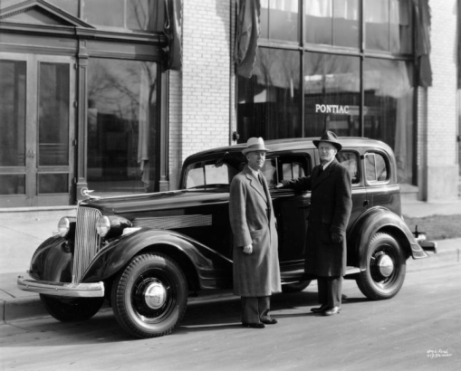 Men pose near an automobile in Denver, Colorado. They wear overcoats and hats. Lettering on a nearby window reads: "Pontiac."