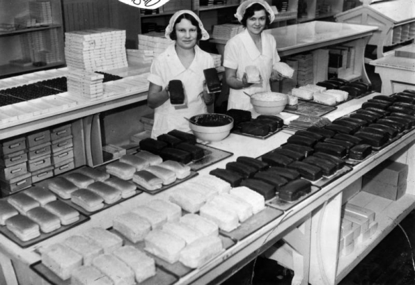 Interior portrait of employees at Mrs. Stover's Candy factory, in Denver, Colorado; women wear caps and pose with cakes and chocolate truffles.