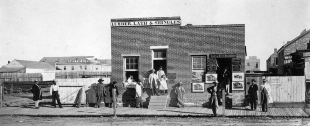 Men and women identified from right to left: "Curtis," "Gestirchild [?]," "Craven," "Hatten," "Perrennoud [?]," "Beers," "Katy Johnson," "Ruten [?]," Aunnell," "Mrs. J. Carr Johnson," "Williams," "McFadden," "McCornie [?], and "J. Carr Johnson," pose outdoors near the Crater Insurance office on Holladay (Market) Street at 16th (Sixteenth) Street in downtown Denver, Colorado. Signs read: "Lumber, Lath, & Shingles," "Life & Fire Insurance," and "Pacific House."