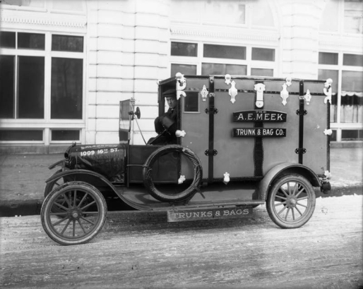 A man sits in the driver's seat of a delivery truck in Denver, Colorado. The rear of the truck is designed to look like a trunk. Signs on the truck read: "A. E. Meek Trunk & Bag Co." and "Trunks & Bags."
