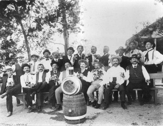 Portrait of Tivoli-Union Brewery employees in Denver, Colorado, posing with beer mugs, kegs, and a pitcher.