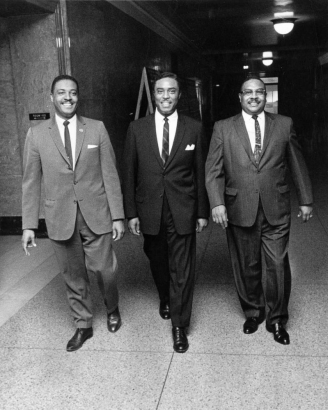 Colorado State Senator George L. Brown, State Representative Daniel Grove, and State Representative Isaac E. Moore (identified left to right), African American men, walk in a hallway in Denver, Colorado. They wear suits and ties.