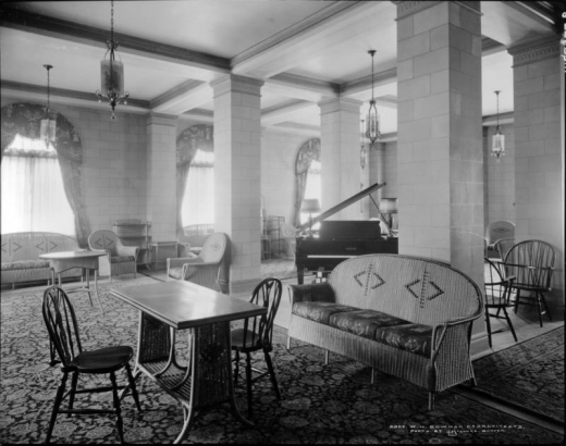 Interior view of lounge, Colburn Hotel, Denver, Colorado; shows  wicker furniture (chairs, table and couch), piano, Oriental rugs, hanging lamps, and brocade window valences and curtains.