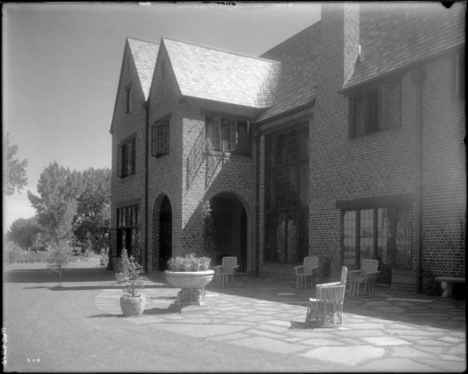 View of Mrs. George W. (Ethel R.) Gano's residence, South University Road, Denver, Colorado, a brick house with arches, picture windows, a flagstone patio, wicker furniture, and potted trees.