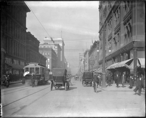 View of 16th (Sixteenth) Street in Denver, Colorado; shows automobiles, a horse-drawn carriage, bicyclists, pedestrians, an electric street car "Union Depot - #244," a clock,  Daniels & Fisher tower construction, and signs: "The Braman Co.," "A. J. Stark & Co.," and "A. T. Lewis & Son."